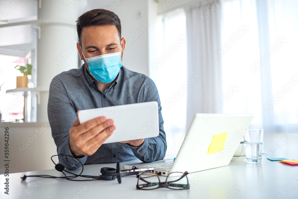 Businessman looking at a tablet PC wearing a healthcare face mask. Man in a medical mask at the office. Businessman with mask using digital tablet at work Businessman looking at a tablet PC wearing a healthcare face mask. Man in a medical mask at the office. Businessman with mask using digital tablet at work
