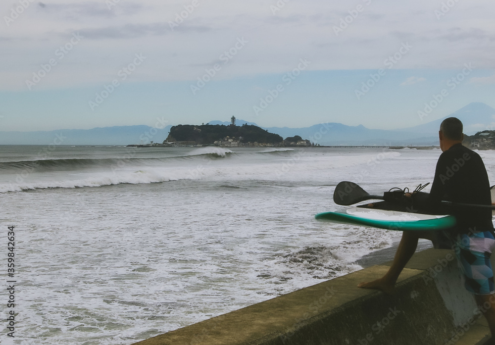 Stand Up Paddle Boarding In Japan near to Enoshima Island with My Fuji in the background. A foreign man with his board looking out to sea where large waves are breaking.