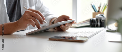 Male designer using digital tablet with stylus pen on white office desk with smartphone, computer device and other supplies
