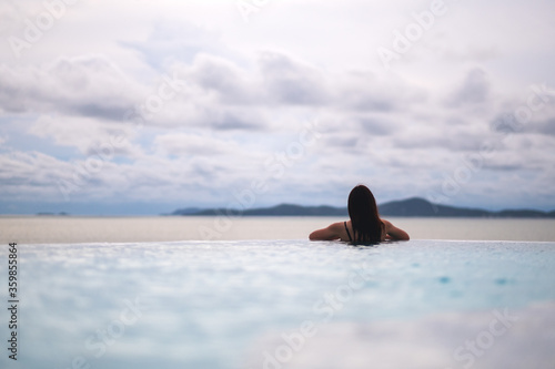 A young asian woman relaxing in infinity swimming pool looking at a beautiful sea view