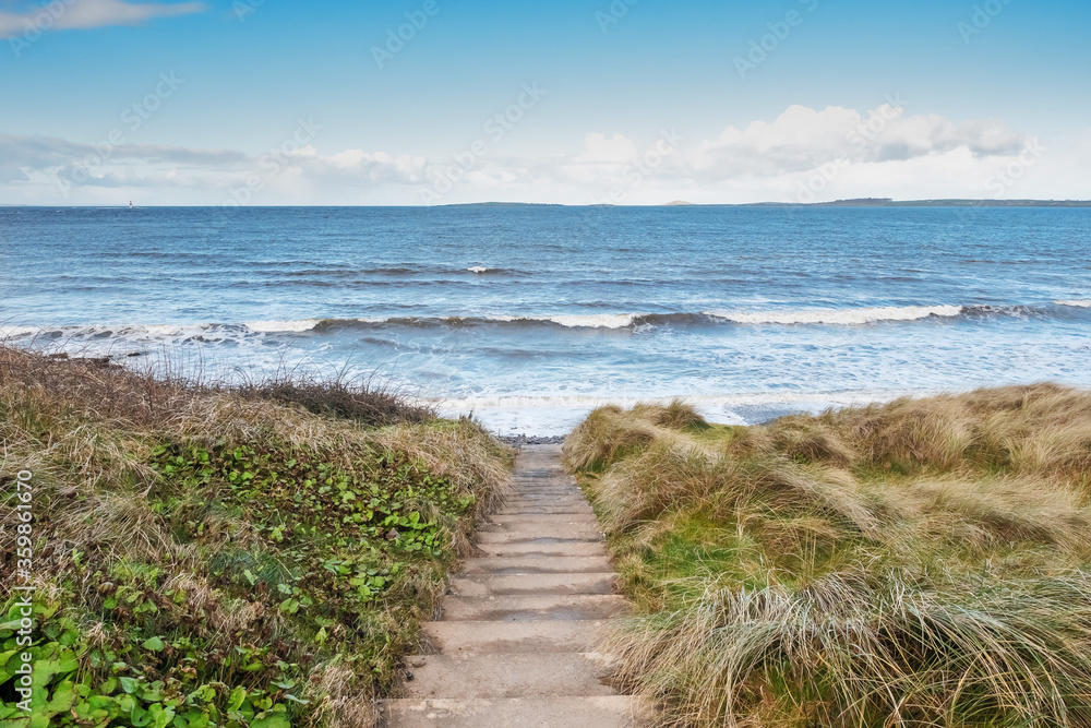 Foot path to the Atlanic ocean, Rosses point, county Sligo Ireland, Blue water surface with waves, clear cloudy sky, Tall grass in foreground. Mountains in the foreground.