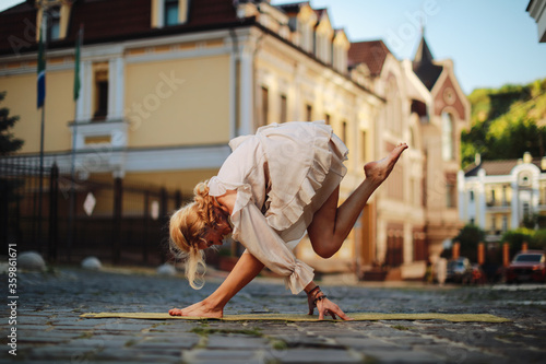 Beautiful blonde woman in nice dress doing a yoga exercise in city on sunset background