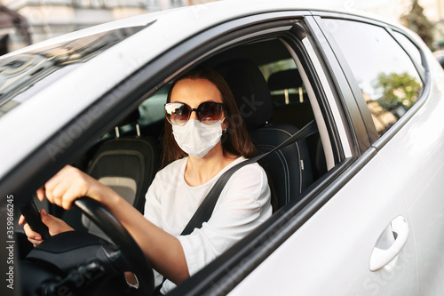 Young woman in sunglasses with protective medical mask driving a car, she holds hands on the steering wheel and looking into camera. Safety during coronavirus pandemic, epidemic covid-19
