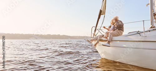 Explore dreams. A happy senior couple sitting on the side of a sail boat on a calm blue sea. Man hugging his woman while enjoying view