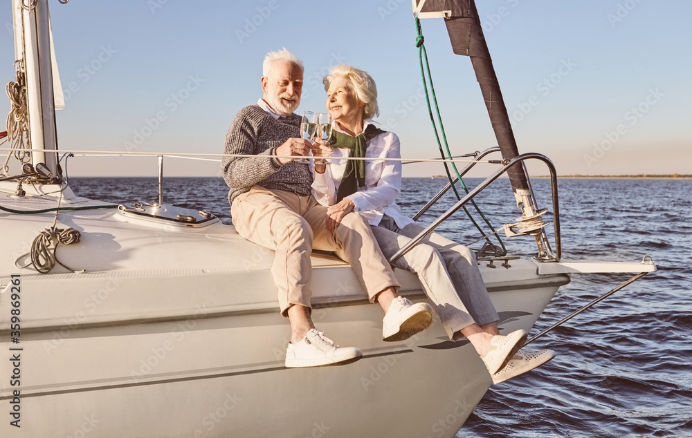 Good vibes. Happy senior couple sitting on the side of sail boat or yacht deck floating in sea. Man and woman drinking wine or champagne, enjoying the view