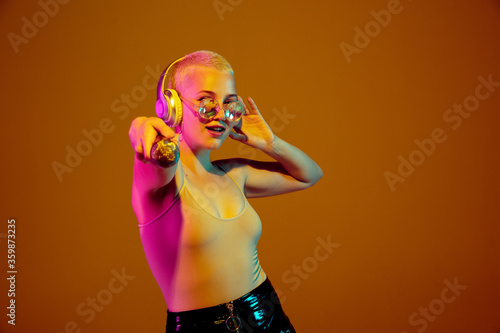 Musician. Young caucasian woman on brown studio background in neon light. Beautiful female model with microphone, speaker. Human emotions, facial expression, sales, ad concept. Freak's culture.