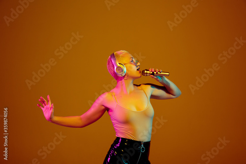 Attractive. Young caucasian woman on brown studio background in neon light. Beautiful female model with microphone, speaker. Human emotions, facial expression, sales, ad concept. Freak's culture.