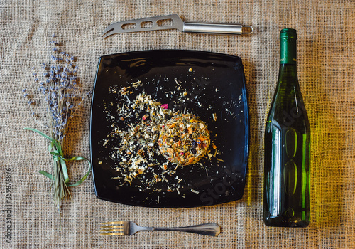 The round head of the French spicy cheese Boulette d'Avesnes sprinkled with different herbs, lies on a black plate. Arrangement: boulette d'avesnes cheese, wine bottle, knife. Film noise