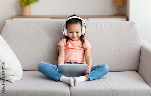 Asian Girl Using Tablet Computer Wearing Wireless Headphones At Home