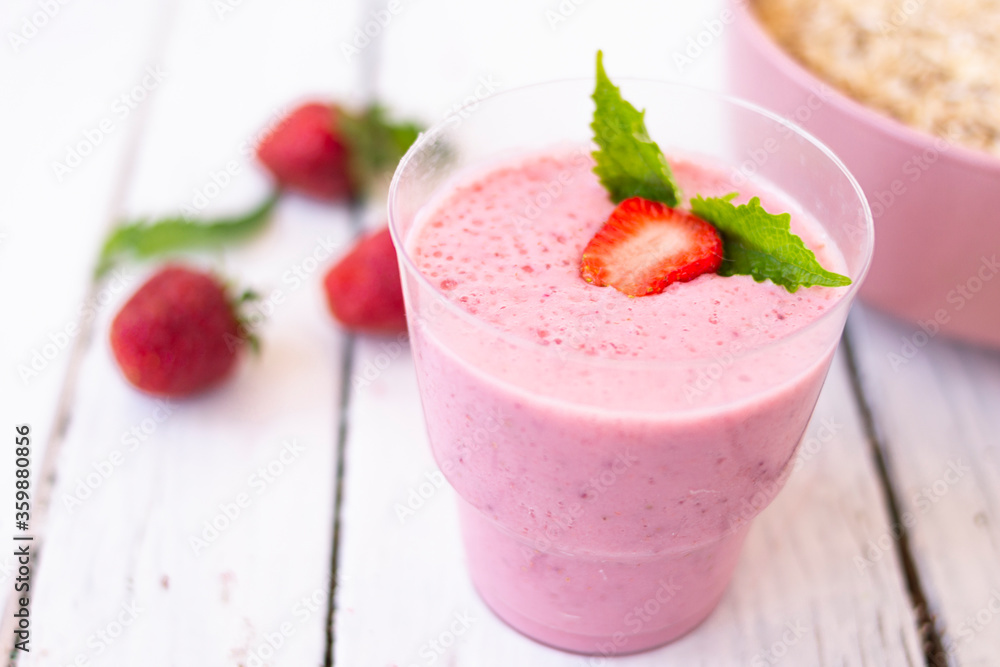 Cocktail with strawberries and milk in a glass decorated with mint and strawberries. Nearby is a pink cup with Hercules flakes, and loose strawberries on a white wooden background. Cocktail with strawberries and milk in a glass decorated with mint and strawberries. Nearby is a pink cup with Hercules flakes, and loose strawberries on a white wooden background.