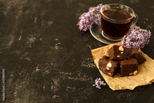 A piece of homemade chocolate cake on crumpled craft paper, with nuts on a black texture table. A cup of tea and lilac flowers decorate a delicious tea party.