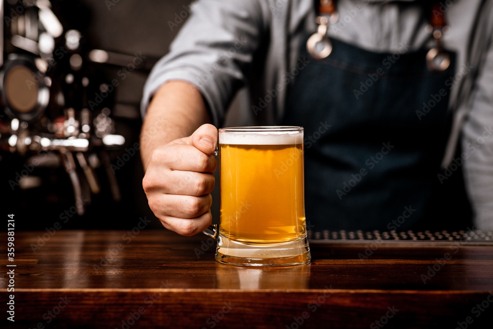 Barman in apron serves beer in glass mug on wooden bar counter