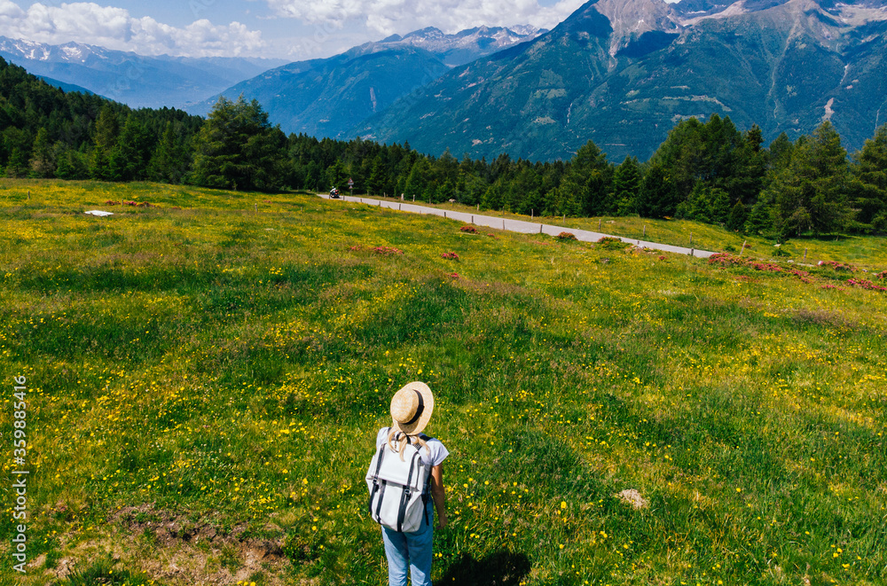 Заголовок: Aerial back view of female tourist in hat admires beautiful meadow on background view of Dolomites mountains.Woman traveler enjoying pure authentic nature while standing near pasture with w Заголовок: Aerial back view of female tourist in hat admires beautiful meadow on background view of Dolomites mountains.Woman traveler enjoying pure authentic nature while standing near pasture with w
