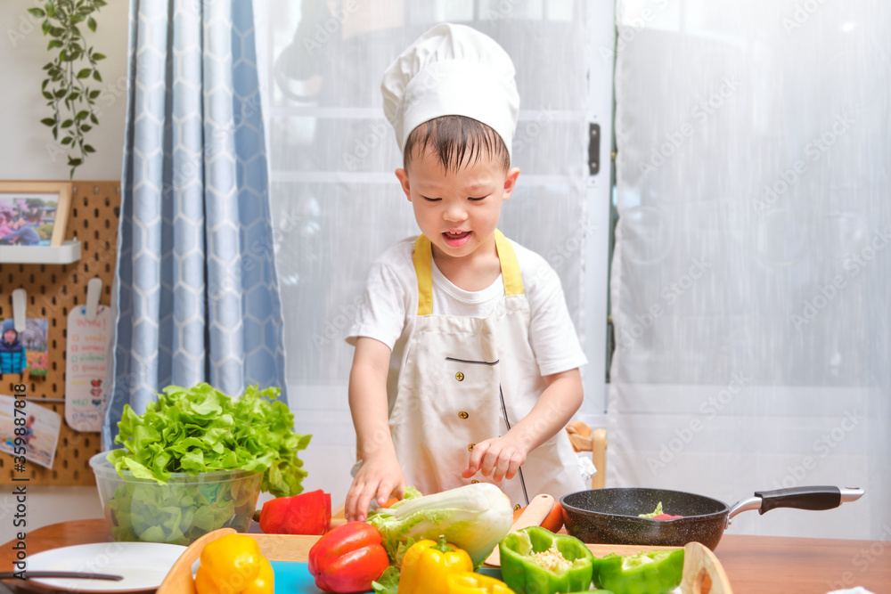 Cute happy smiling Asian little boy child wearing chef hat and apron having fun preparing, cooking healthy food in kitchen, Fun indoor activities for kindergarten kids, photo in real life interior