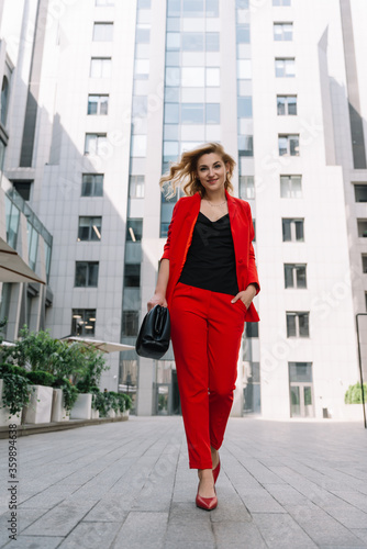Young business woman blonde in a red suit is walking outdoors in the courtyard of a business center. Stylish people. Soft selective focus.
