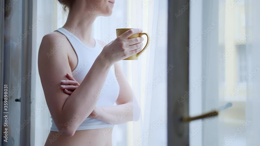 Young woman standing in window at home daydreaming and drinking morning coffee. Unrecognizable person. High quality 4K video footage. Young woman standing in window at home daydreaming and drinking morning coffee. Unrecognizable person. High quality 4K video footage.