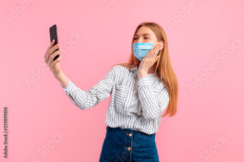 Happy girl in a medical protective mask on her face, taking a selfie on a mobile phone, on an pink background. coronavirus