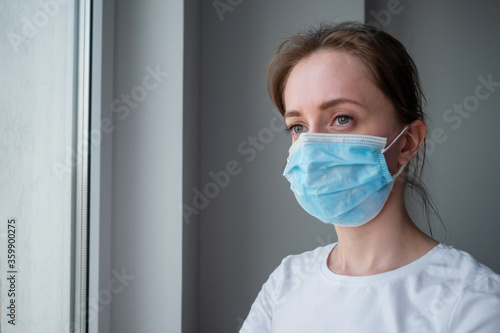 Portrait of pensive woman wearing medical face mask and looking out of window in room with grey wall at home. Self isolation, prevention, quarantine, COVID-19, coronavirus, safety concept