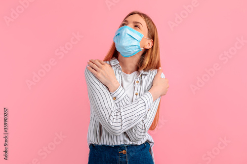 young woman in a medical protective mask holding herself in an embrace, against an isolated pink background. Love yourself