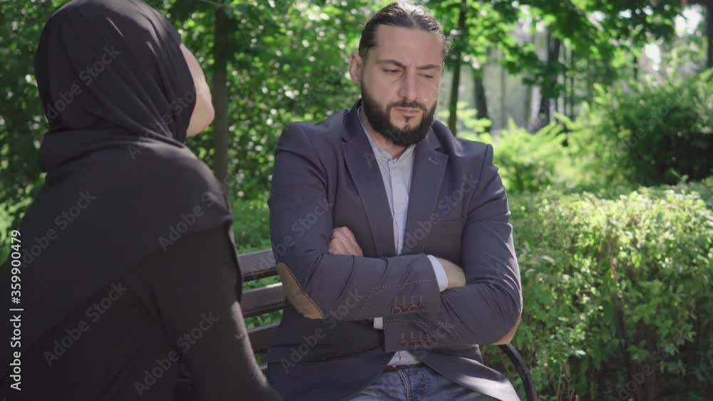 Proud bearded Middle Eastern man sitting on bench with hands crossed as unrecognizable woman in hijab talking to him. Portrait of argued Middle Eastern couple in park on sunny summer day. Conflict. Proud bearded Middle Eastern man sitting on bench with hands crossed as unrecognizable woman in hijab talking to him. Portrait of argued Middle Eastern couple in park on sunny summer day. Conflict.