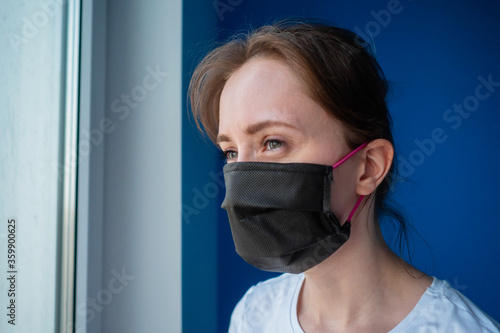 Portrait of pensive woman wearing black medical face mask and looking out of window in room with blue wall at home. Self isolation, prevention, quarantine, COVID-19, coronavirus, safety concept