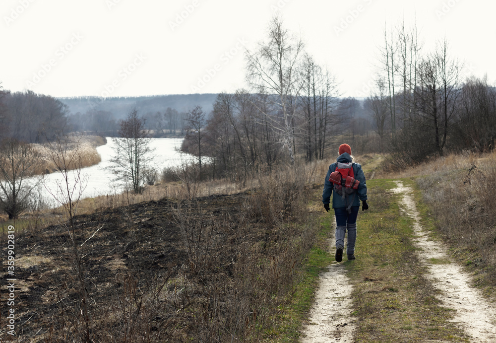 A girl in an autumn jacket, hat and with a backpack takes a walk on nature in early spring. He looks at the vast expanses and enjoys the view.