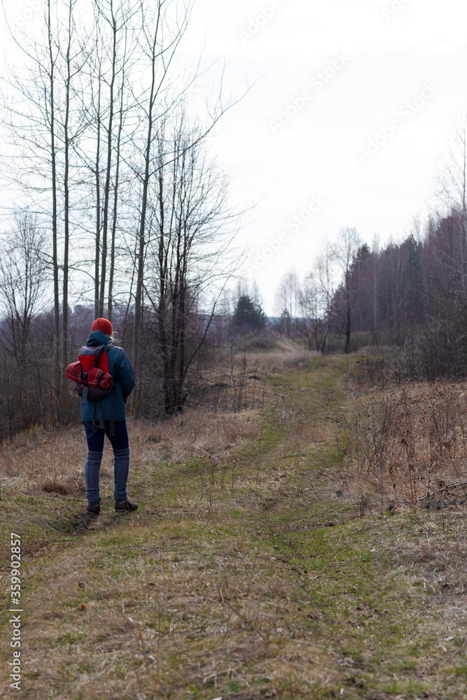 A girl in an autumn jacket, hat and with a backpack takes a walk on nature in early spring. He looks at the vast expanses and enjoys the view.