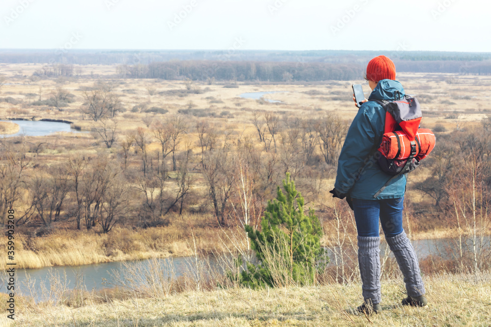 A girl in an autumn jacket, hat and with a backpack takes a walk on nature in early spring. He looks at the vast expanses and enjoys the view.