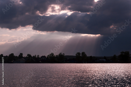 Dark purple clouds at sunset before a thunderstorm. Reflection of the sky in the water. Beautiful evening landscape. Pond, lake. Sun rays through thick clouds. © Anastasiya Gracheva