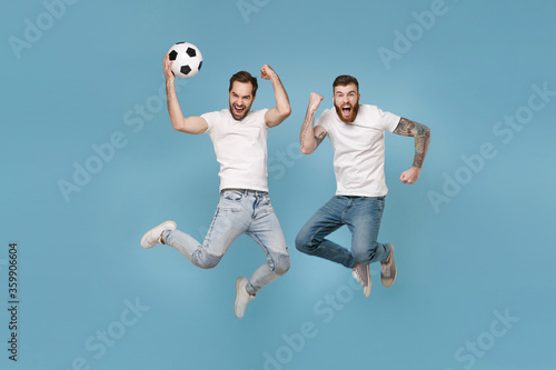 Crazy men guys friends in white t-shirt isolated on pastel blue background. Sport leisure lifestyle concept. Cheer up support favorite team with soccer ball, jumping, doing winner gesture, screaming.