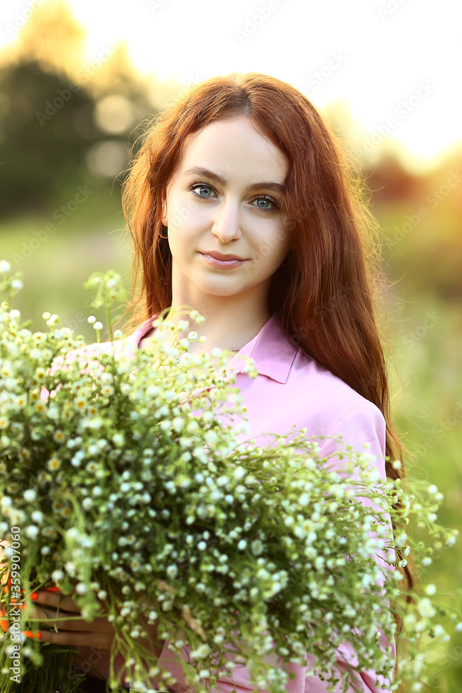 A young beautiful red-haired girl close-up is looking at the camera with a huge bouquet of field daisies in a pink dress. Sunset on a summer evening. Effect bokeh, flash. A young beautiful red-haired girl close-up is looking at the camera with a huge bouquet of field daisies in a pink dress. Sunset on a summer evening. Effect bokeh, flash.