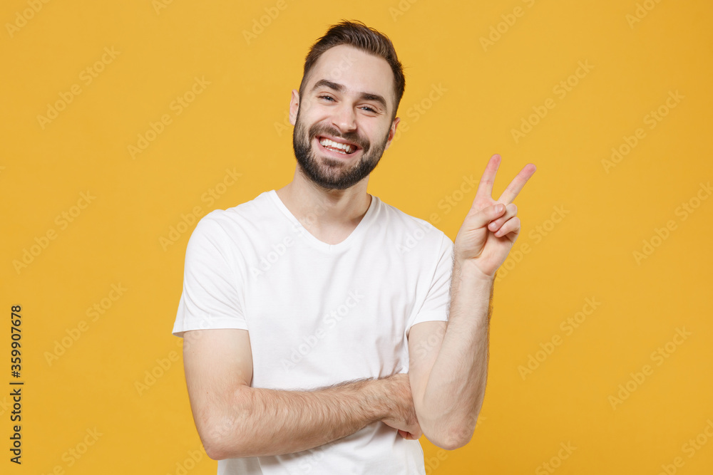 Smiling young bearded man guy in white casual t-shirt posing isolated on yellow wall background studio portrait. People sincere emotions lifestyle concept. Mock up copy space. Showing victory sign. Smiling young bearded man guy in white casual t-shirt posing isolated on yellow wall background studio portrait. People sincere emotions lifestyle concept. Mock up copy space. Showing victory sign.