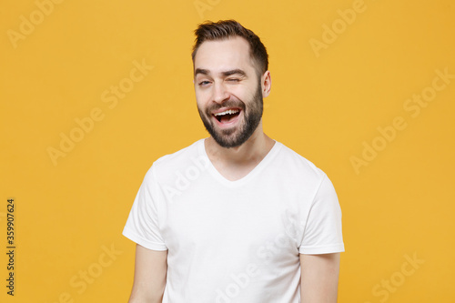 Cheerful funny young bearded man guy 20s in white casual t-shirt posing isolated on yellow wall background studio portrait. People sincere emotions lifestyle concept. Mock up copy space. Blinking.
