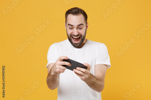 Excited young bearded man guy in white casual t-shirt posing isolated on yellow wall background studio portrait. People lifestyle concept. Mock up copy space. Play game with mobile phone, screaming.