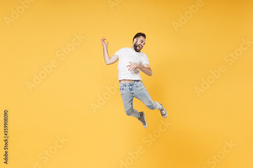 Funny young bearded man guy in white casual t-shirt posing isolated on yellow background studio portrait. People emotions lifestyle concept. Mock up copy space. Jumping hold hands like playing guitar.