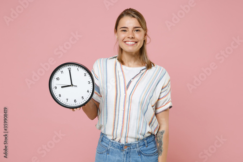 Smiling funny young blonde woman girl in casual striped shirt posing isolated on pastel pink wall background studio portrait. People sincere emotions lifestyle concept. Mock up copy space. Hold clock.