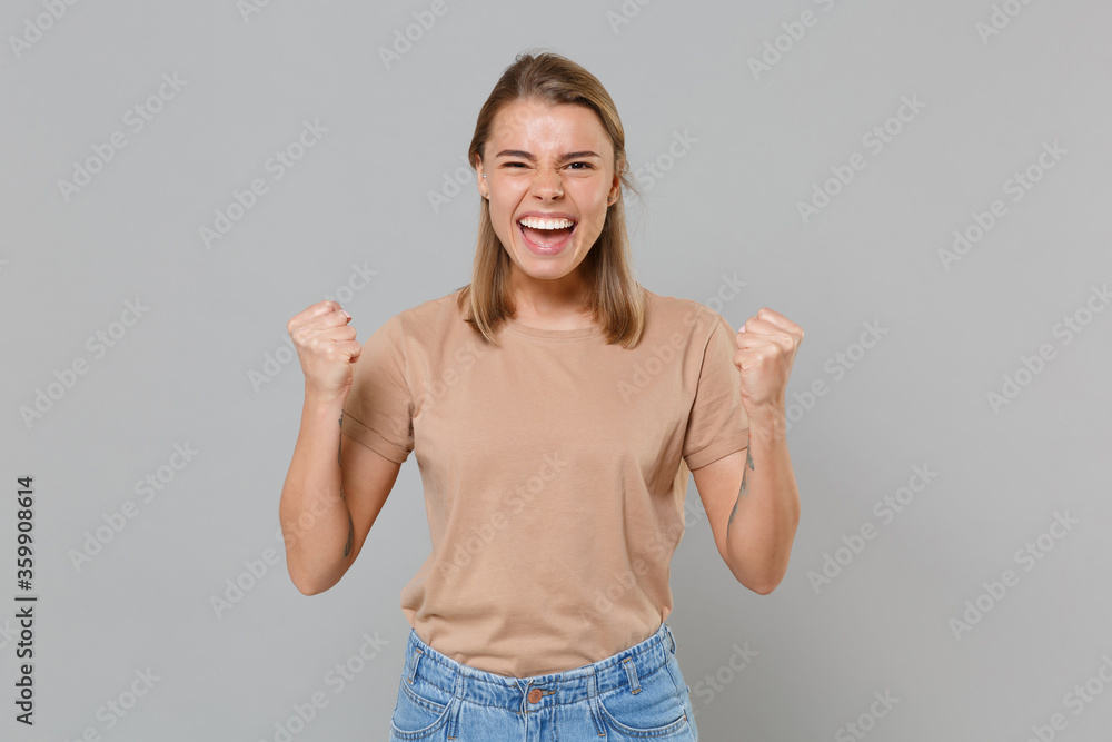 Happy young blonde woman girl in casual beige t-shirt posing isolated on gray wall background studio portrait. People sincere emotions lifestyle concept. Mock up copy space. Doing winner gesture. Happy young blonde woman girl in casual beige t-shirt posing isolated on gray wall background studio portrait. People sincere emotions lifestyle concept. Mock up copy space. Doing winner gesture.