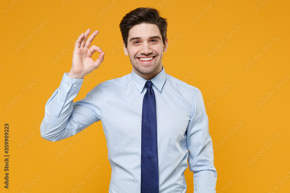 Smiling young business man in classic blue shirt tie posing isolated on yellow wall background studio portrait. Achievement career wealth business concept. Mock up copy space. Showing OK gesture. Smiling young business man in classic blue shirt tie posing isolated on yellow wall background studio portrait. Achievement career wealth business concept. Mock up copy space. Showing OK gesture.