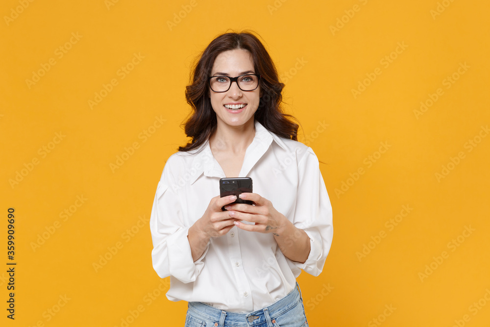 Smiling young brunette business woman in white shirt glasses isolated on yellow wall background . Achievement career wealth business concept. Mock up copy space. Using mobile phone typing sms message.