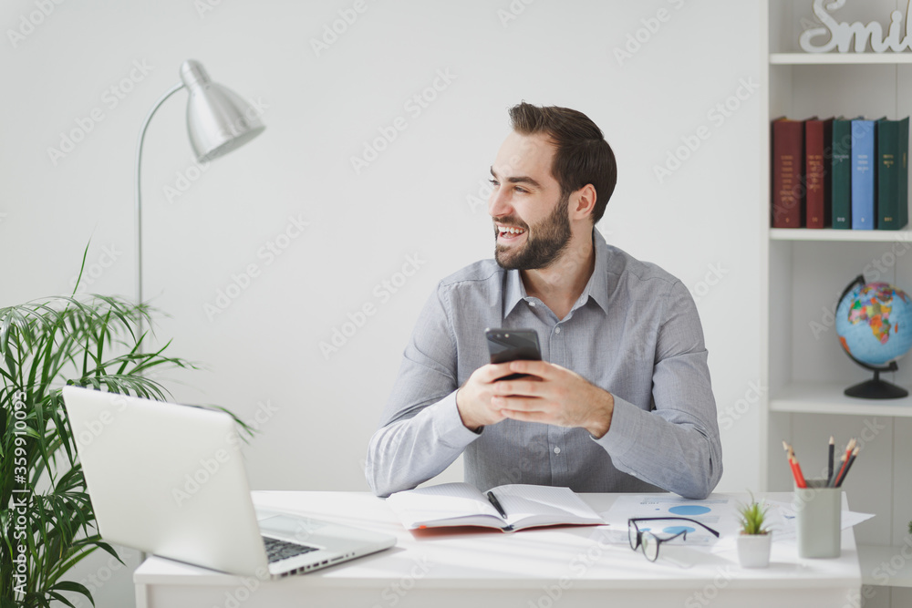 Cheerful young business man in gray shirt sitting at desk work on laptop pc computer in light office on white wall background. Achievement business career concept. Using mobile phone, looking aside.