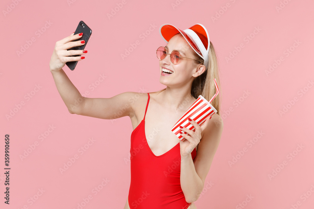 Smiling young woman girl in red one-piece swimsuit cap glasses isolated on pink background. People summer vacation rest lifestyle concept. Doing selfie shot on mobile phone, hold cup of cola or soda. Smiling young woman girl in red one-piece swimsuit cap glasses isolated on pink background. People summer vacation rest lifestyle concept. Doing selfie shot on mobile phone, hold cup of cola or soda.