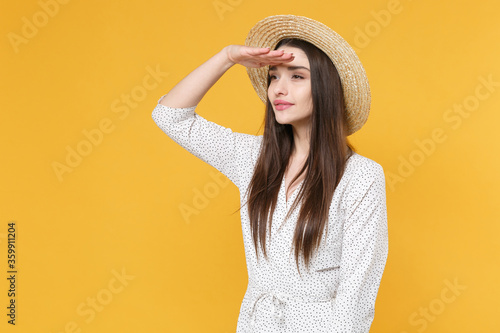 Beautiful young brunette woman girl in white dress hat posing isolated on yellow background in studio. People lifestyle concept. Mock up copy space. Holding hand at forehead looking far away distance.