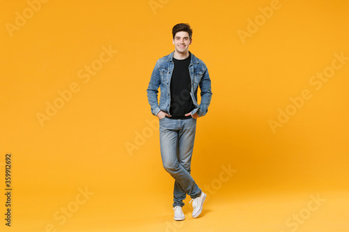 Smiling young man guy 20s wearing casual denim clothes posing isolated on yellow background studio portrait. People sincere emotions lifestyle concept. Mock up copy space. Holding hands in pockets.