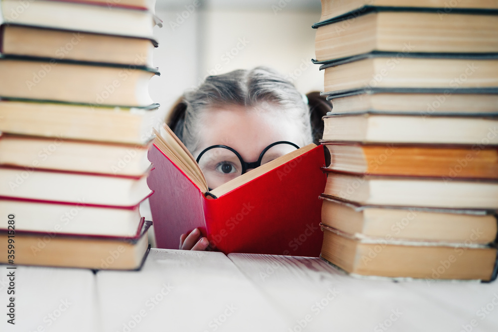 Serious little girl in round shaped glasses practicing reading between books pile. Primary education concept. Serious little girl in round shaped glasses practicing reading between books pile. Primary education concept.