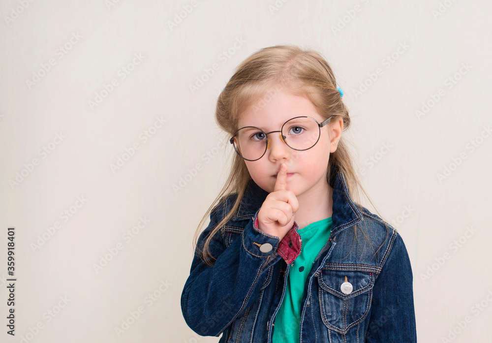 Little girl in eyeglasses putting finger up to lips and ask silence. Child doing a Please Keep Quiet gesture towards the camera. Little girl in eyeglasses putting finger up to lips and ask silence. Child doing a Please Keep Quiet gesture towards the camera.