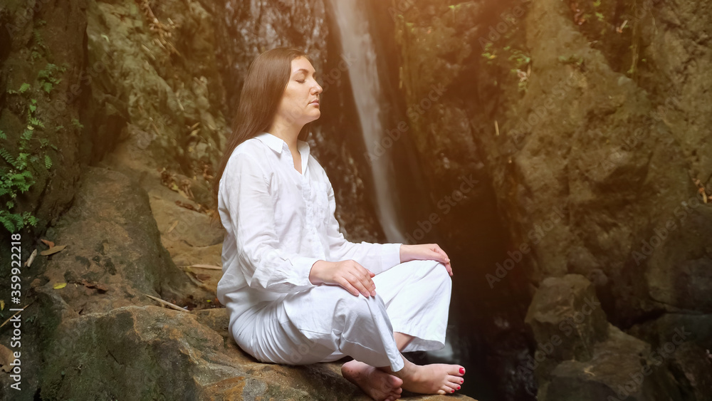 concentrated woman with loose dark hair in white costume meditates sitting in yoga lotus position on rock against tropical waterfall closeup concentrated woman with loose dark hair in white costume meditates sitting in yoga lotus position on rock against tropical waterfall closeup