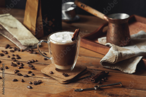 glass transparent coffee Cup with double walls with coffee poured in layers with cinnamon and milk foam on rough wooden table and scattered coffee beans, craft bags and Turkish coffee pot behind photo