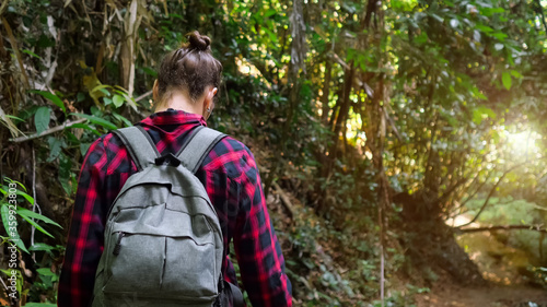 woman with hair bun wearing checkered shirt and backpack enjoys hiking and walks in lush tropical forest close backside view