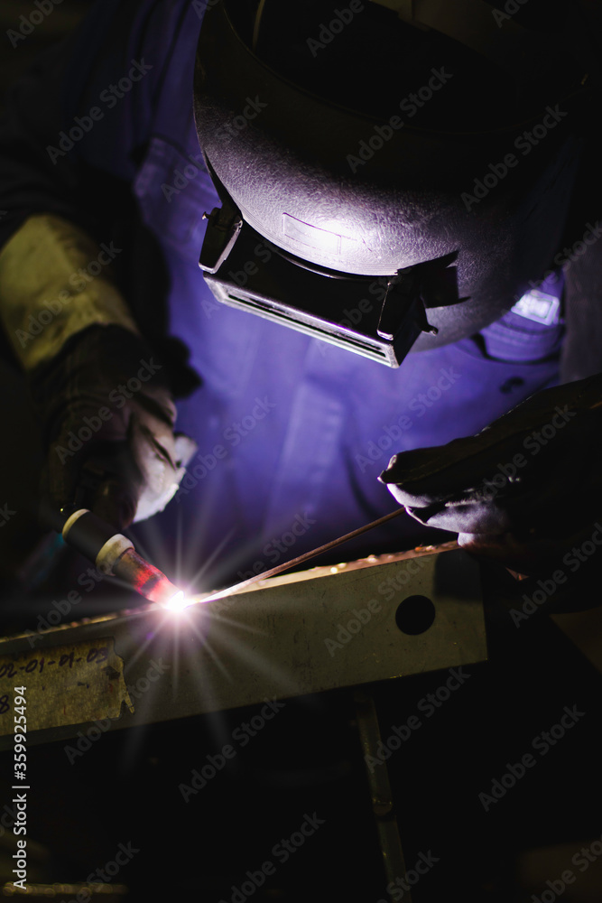 Welder is welding the steel in the factory. Weld the steel in dark. Workers wearing industrial uniforms and welding masks at a welding factory protect from welding sparks. Occupational safety concept Welder is welding the steel in the factory. Weld the steel in dark. Workers wearing industrial uniforms and welding masks at a welding factory protect from welding sparks. Occupational safety concept