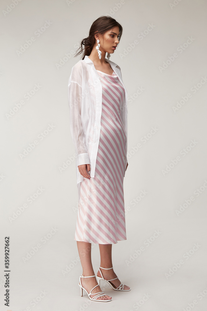 Sexy fashion model beautiful woman pretty face makeup, tanned skin wear summer collection clothes style for romantic date walk trendy light silk in white pink stripes long dress white shirt earrings. Sexy fashion model beautiful woman pretty face makeup, tanned skin wear summer collection clothes style for romantic date walk trendy light silk in white pink stripes long dress white shirt earrings.